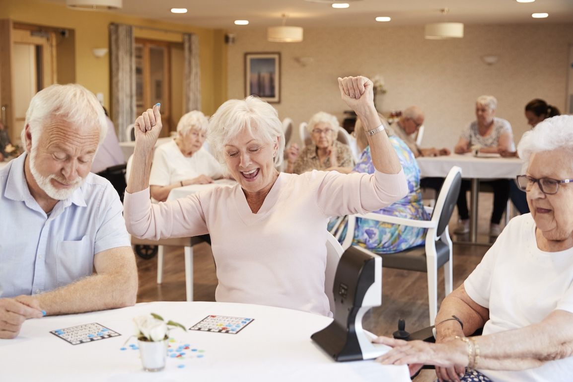 Senior adult woman joyfully raising arms while playing bingo at a senior living community, surrounded by peers enjoying the game.