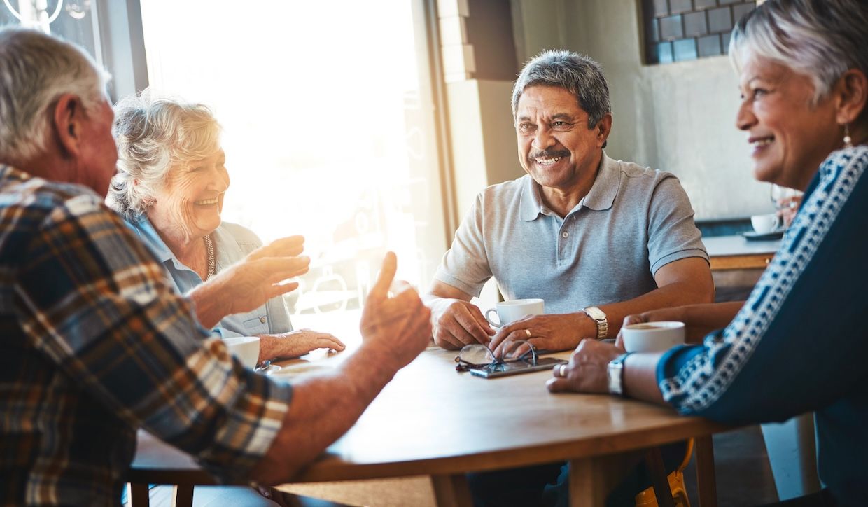 Group of senior adults sitting together at a table, laughing and talking in a bright, social setting at a senior living community.
