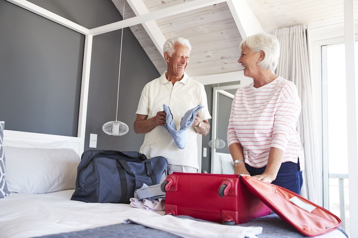 Senior couple smiling while packing a suitcase in a cozy bedroom, preparing for vacation travel.