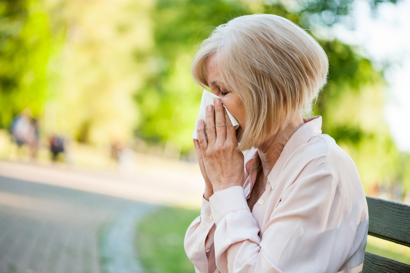 Adult woman is sitting in park and blowing nose. She is experiencing seasonal allergies.