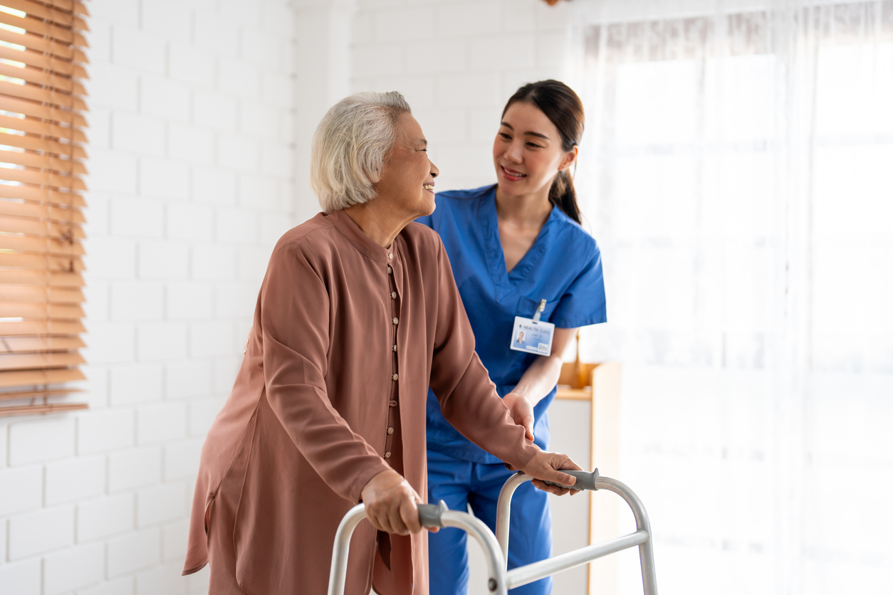 Senior woman using a walker with the assistance of a smiling healthcare professional in blue scrubs at an assisted living facility