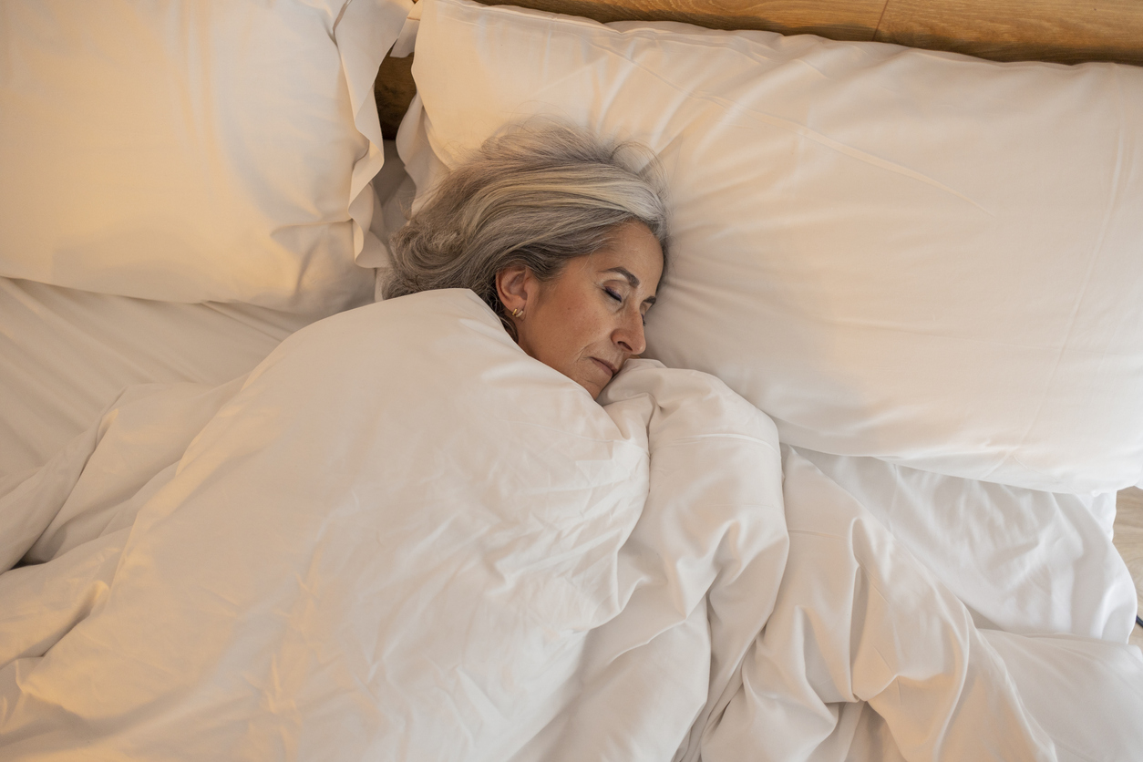 Senior adult woman peacefully asleep in a cozy bed with white pillows and blankets.
