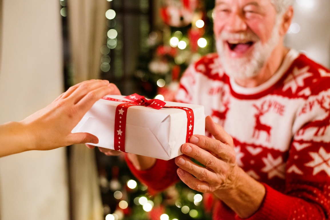 Senior man in a festive sweater receiving a wrapped Christmas gift in front of a decorated holiday tree.