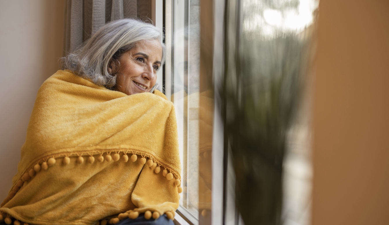 Senior woman wrapped in a warm blanket looking out the window on a winter day, representing cozy indoor living and seasonal wellness for older adults.